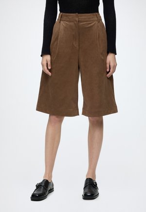 Persona que lleva pantalones cortos marrones de pierna ancha hasta la rodilla, top negro de manga larga y mocasines de cuero negro, de pie frente a un fondo liso.