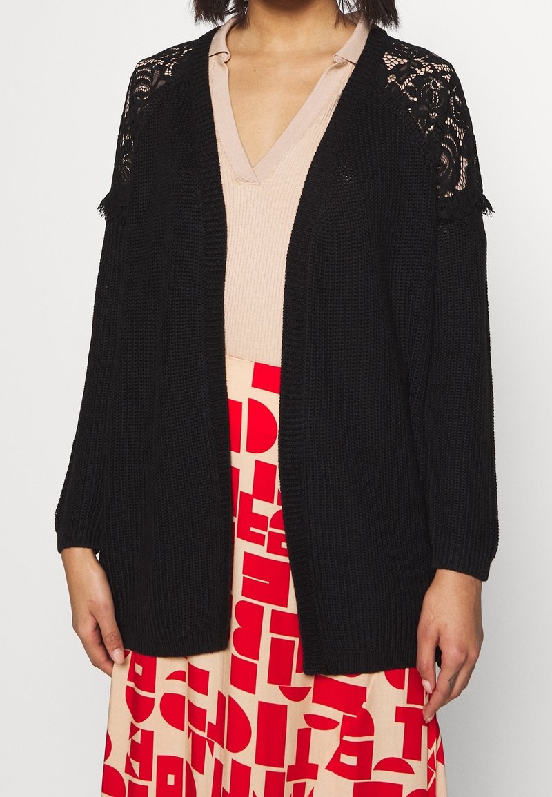 Schwarze Strickjacke mit offenem Vorderteil, Spitzenbesatz an den Schultern und Dreiviertelärmeln. Getragen über einem hellen V-Ausschnitt-Oberteil und einem gemusterten Rock.