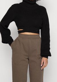 En person bär en svart kortärmad polotröja med utskärningar, tillsammans med bruna sweatpants med en broderad logotyp.