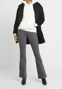 Černý strukturovaný kabát, bílá mikina s kapucí a šňůrkami, šedé zvonové kalhoty a černé špičaté kotníkové boty. Tento outfit spojuje casual a chic prvky.