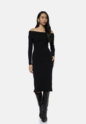 Vrouw die een zwarte off-shoulder midi-jurk met lange mouwen draagt, gecombineerd met zwarte leren kniehoge laarzen op een neutrale achtergrond.