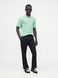 Homme portant un polo vert menthe, un pantalon foncé, une ceinture marron et des chaussures de ville beige et marron, debout devant un fond blanc.