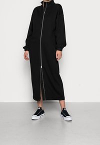 Sort lynlås kjole lavet af blødt stof, med lange ærmer og høj krave. Model stylet med sorte sneakers for et sporty udseende.