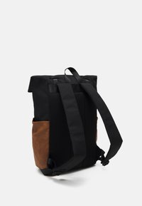 Sac à dos en tissu noir avec des accents en cuir brun, détails en maille, bretelles réglables et une poignée supérieure. Poche latérale visible.