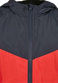 Veste rouge et marine avec capuche, dotée d'une fermeture éclair centrale et d'un design en chevrons. Fabriquée en tissu lisse et léger.