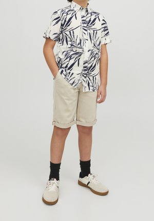 Koszula z krótkim rękawem w biało-granatowy wzór liści, zestawiona z beżowymi szortami z podwiniętymi nogawkami. Czarne skarpetki i beżowe trampki dopełniają wygląd.