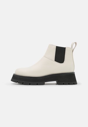 Bota Chelsea de cuero color crema con paneles laterales elásticos negros, suela gruesa con tacos y lengüeta en el talón para facilitar su colocación.