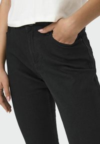 Crne traperice od denima sa slim fit krojem, s prednjim džepovima, suptilnim detaljima šivanja i visokim strukom. Uključuju zatvaranje na dugme.