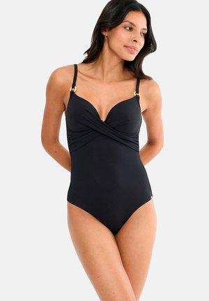 Femme portant un maillot de bain une-pièce noir avec un détail en tissu croisé et des bretelles ajustables, debout les mains derrière le dos.
