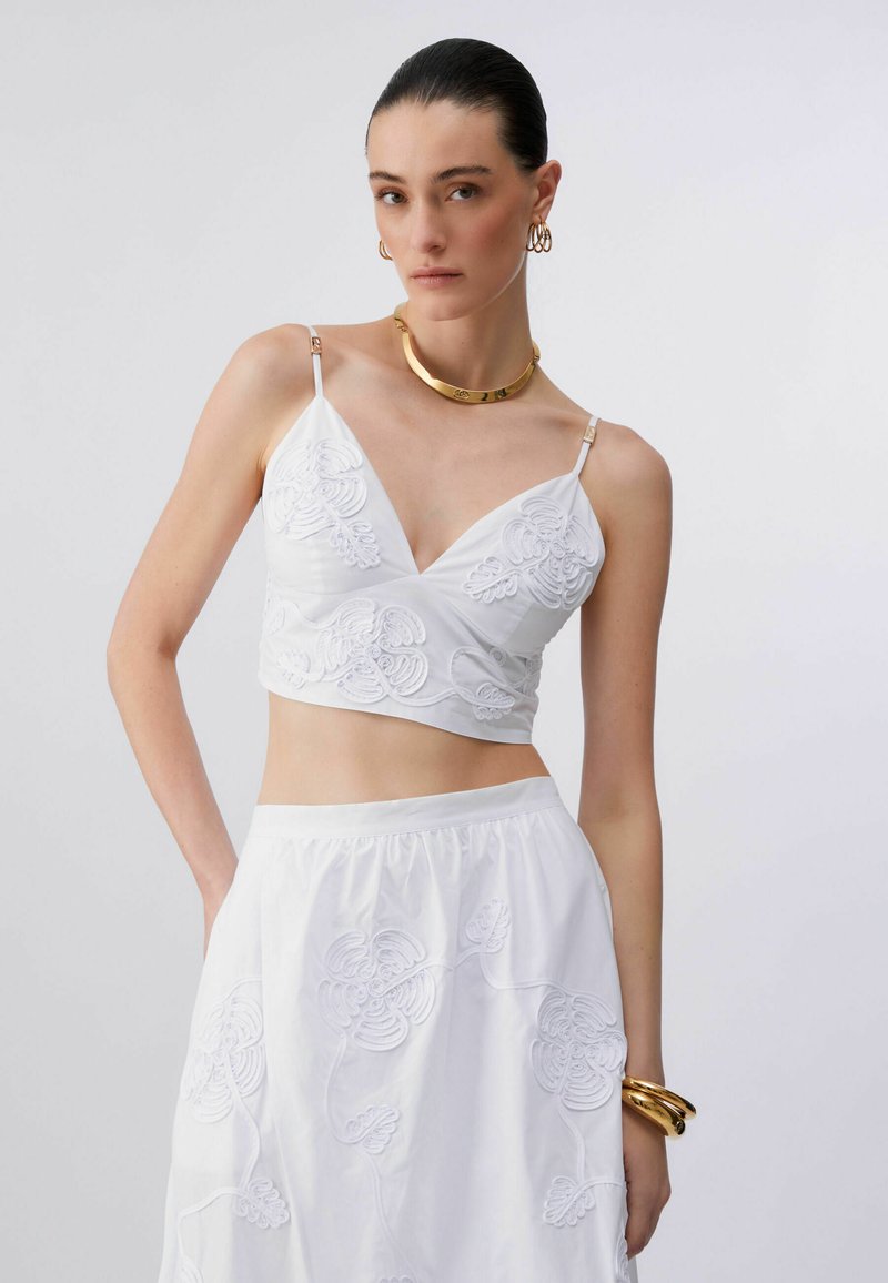 Femme portant un ensemble crop top brodé blanc et jupe, avec un collier en or, des boucles d'oreilles créoles et des bracelets, devant un fond uni.