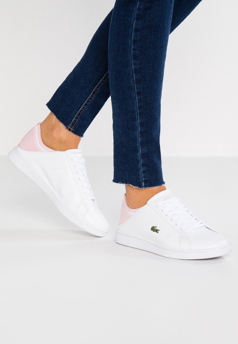Personne portant des baskets Lacoste blanches et roses avec un jean bleu foncé sur un fond blanc uni.