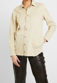 Beige knapp-up skjorta i bomull, med två framfickor, fransad fåll och en subtil broderad detalj på ärmen.