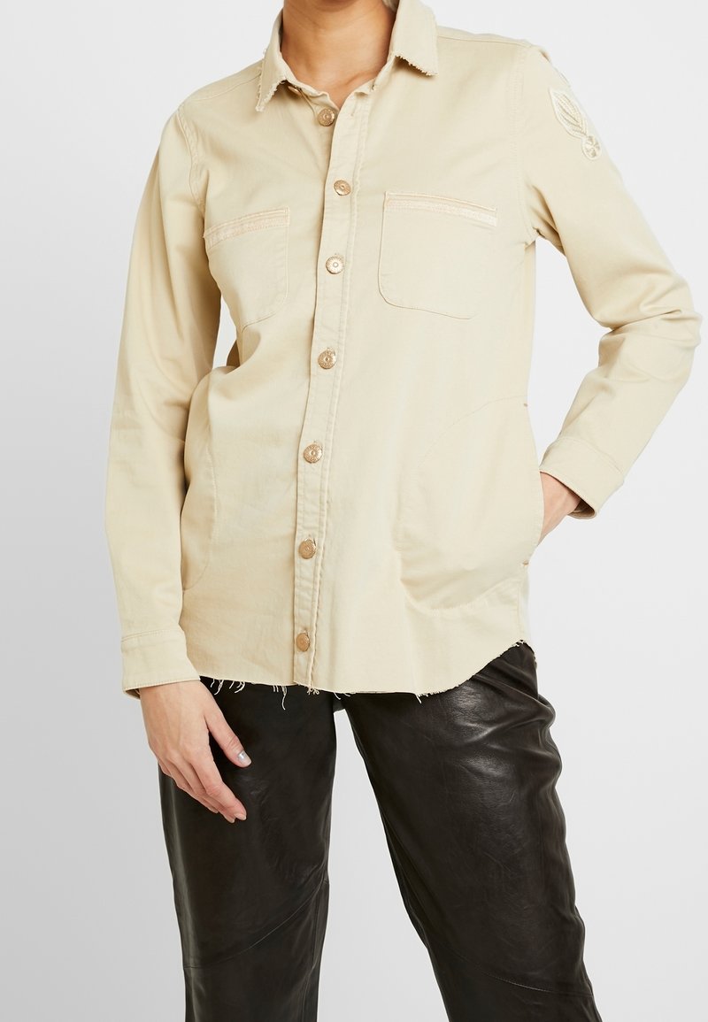 Beige knapp-up skjorta i bomull, med två framfickor, fransad fåll och en subtil broderad detalj på ärmen.