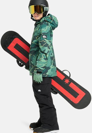 Zielona kurtka snowboardowa w kamuflażu, czarne spodnie narciarskie, rękawice i kask. Deska snowboardowa ma czerwony wzór na czarnym tle. Duże lustrzane gogle.