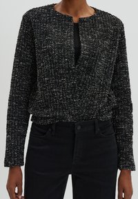 Veste longue à manches longues texturée noire avec une coupe ample et une ouverture sur le devant, présentant un motif tissé subtil et un col rond. Associée à un jean noir.