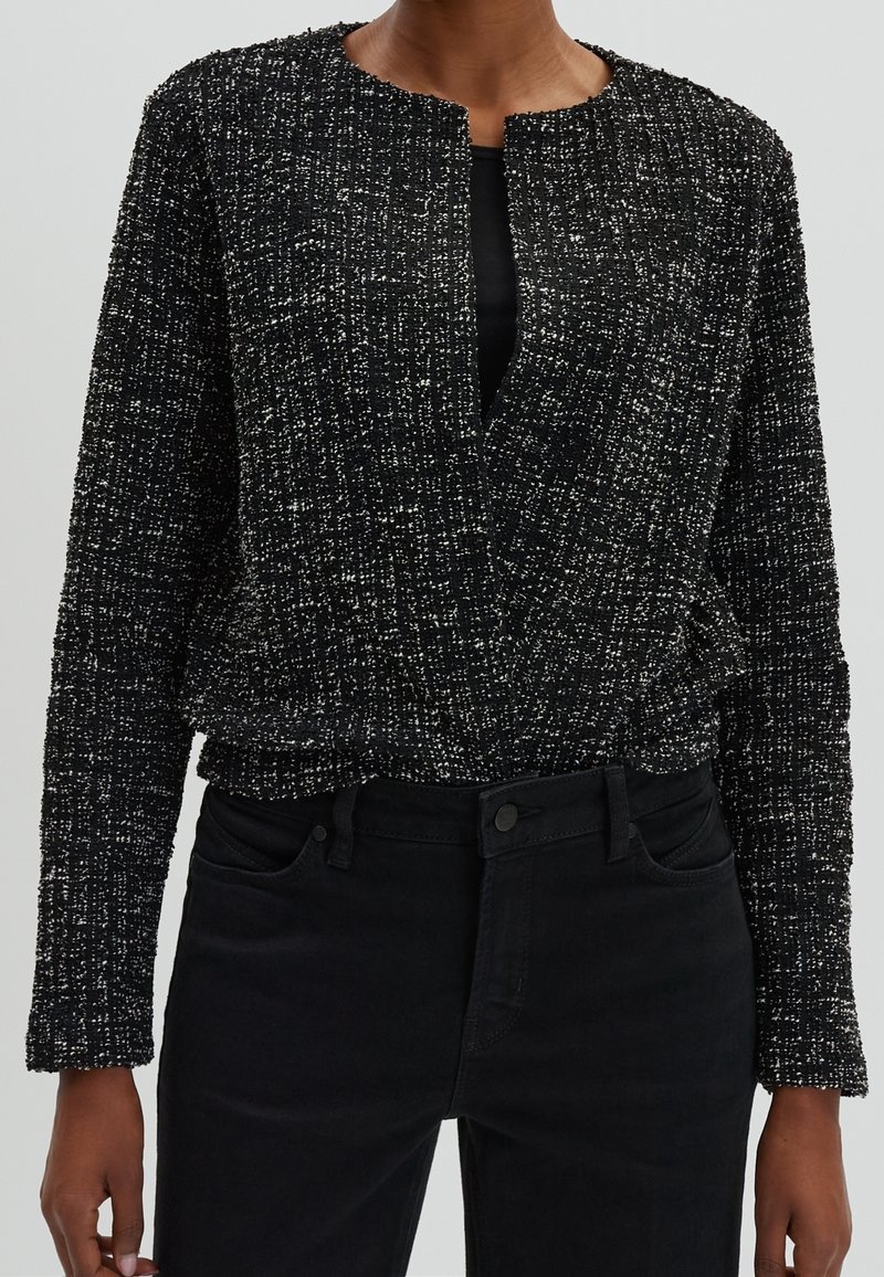 Veste longue à manches longues texturée noire avec une coupe ample et une ouverture sur le devant, présentant un motif tissé subtil et un col rond. Associée à un jean noir.