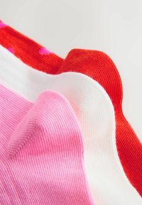 Trīs riboti adīti zeķes rozā, baltā un sarkanā krāsā, novietotas blakus uz gaiša fona.