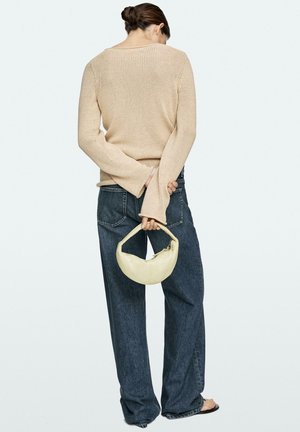 Persoon draagt een beige gebreide trui en losse donkere jeans, houdt een kleine crèmekleurige handtas achter de rug vast, met de rug naar de kijker toe.