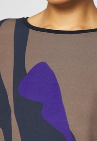 Vícebarevný top z lehké látky. Obsahuje abstraktní vzory v hnědé, námořnické a fialové barvě. Kulatý výstřih se pevným černým lemem.