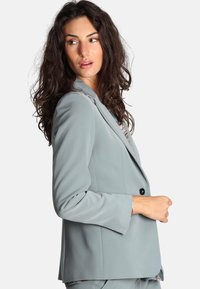Blazer su misura blu chiaro con rever, chiusura con un solo bottone e maniche lunghe. Tessuto liscio e silhouette aderente.