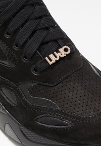Sneaker in suede nero con perforazioni, spesse laccetti neri e un charm decorativo in tonalità oro "LIU JO" sui lacci.