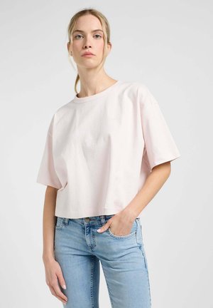 T-shirt corta in cotone rosa con colletto rotondo e maniche corte, caratterizzata da un sottile dettaglio del logo sul petto. Abbinata a jeans azzurri chiari.