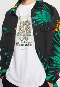 Czarna kurtka z polaru z zielonymi i czerwonymi wzorami kwiatowymi; nałożona na białą koszulkę z kolorową abstrakcyjną grafiką i logo.