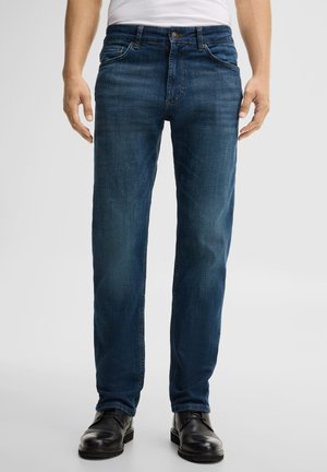 Mann trägt dunkelblaue Jeans mit geradem Bein, schwarze elegante Schuhe und ein weißes Hemd vor einem einfarbigen hellen Hintergrund.