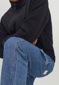 Zwarte fleecejas met ritssluiting en hoge kraag, gecombineerd met blauwe spijkerbroeken met een klassieke snit en subtiele textuurdetails.