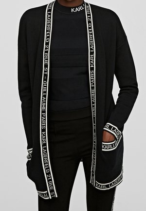 Schwarzer langer Cardigan mit weißem Textbesatz mit der Aufschrift "Karl Lagerfeld 21 Rue St Guillaume Paris", getragen über einem schwarzen Top und Hose.