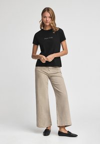 Camiseta de algodón negra con el logo "Polo Club" en blanco, combinada con pantalones de pierna ancha en beige claro y mocasines negros. Fondo neutro.