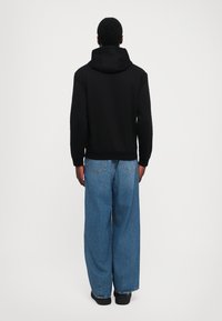 Felpa nera con cappuccio, dal taglio rilassato e polsini a costine, abbinata a jeans in denim chiaro e larghi. I jeans presentano tasche posteriori visibili.
