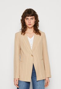 Blazer a righe beige con chiusura a un bottone, due tasche anteriori e vestibilità sartoriale. Indossato sopra una maglietta bianca a scollo a V e jeans.