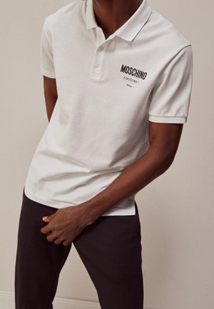 Mann trägt weißes Moschino Couture Polohemd und dunkle Hose, mit einer Hand in Taillennähe vor einfarbigem Hintergrund.