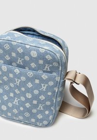 Denim crossbody taška vo svetlom modrom odtieni s bielymi monogramovými vzormi. Disponuje horným zipsom a béžovým nastaviteľným popruhom. Kompaktný obdĺžnikový tvar.