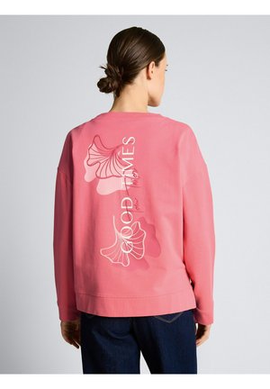 Kobieta z ciemnym kokiem, ubrana w różową bluzę z białą kwiatową grafiką i napisem "Good Times" na plecach, w połączeniu z ciemnymi dżinsami.