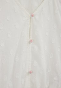 Biela bavlnená blúzka s textúrovaným bodkovaným vzorom a výstrihom do V. Pät ružových gombíkov s kvetinovým motívom slúži ako zapínanie na prednej strane.
