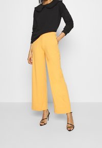 Top nero a maniche lunghe con colletto arricciato, abbinato a pantaloni gialli a gamba larga e sandali neri con cinturini. Design semplice, texture morbida.