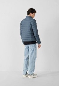 Grijze pufferjack met horizontaal geblokte design, hoge kraag en een gladde textuur. Draagt over een zwarte top, gecombineerd met lichtblauwe jeans.