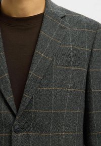 Tumši pelēks žakete ar houndstooth rakstu un gaismas brūnas akcentiem. Divas pogas priekšpusē, ar iegrieztām atlokiem, vilnas audums ar struktūru. Zem tā vienkārša brūna krekla.