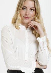 Bílá halenka s dlouhými rukávy, volánkovým lemem a manžetami, na zapínání s knoflíky a vyrobená z hladkého, lehkého materiálu.