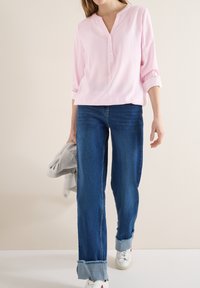 Femme portant un chemisier rayé rose, manches retroussées, jean bleu remonté, baskets blanches, tenant une veste gris clair dans une main.