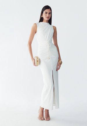Frau in ärmellosem weißem gerafftem Maxikleid mit vorderem Schlitz, hält eine beige Clutch, trägt goldene Ohrringe, Armbänder und Riemchenabsätze.