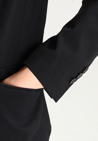 Czarny, dopasowany rękaw marynarki z gładkiej tkaniny, z trzema czarnymi guzikami przy mankiecie oraz widoczną boczną kieszenią przy dole.