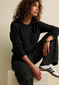 Maglione nero a maglia con collo a coste, pantaloni a palazzo grigi a quadri e sneaker bianche con strisce nere. Orologio dorato al polso.