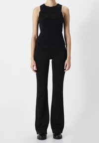 Kobieta w czarnej bluzce bez rękawów z koronkowym wykończeniem oraz czarnych spódnicach w kształcie dzwonu, stojąca na białym tle.