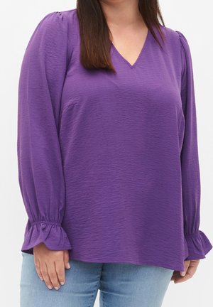 Femme portant un chemisier violet texturé à manches longues avec des poignets volantés et un jean bleu clair, sur un fond uni.