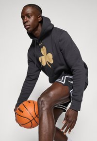 Felpa nera con un grande design di trifoglio dorato, abbinata a pantaloncini sportivi neri e bianchi. Il soggetto tiene in mano un pallone da basket arancione.