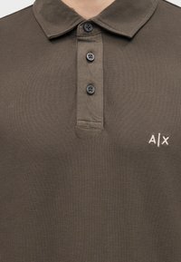Polo marron texturé avec trois boutons noirs et logo "A|X" blanc brodé sur le côté gauche de la poitrine.