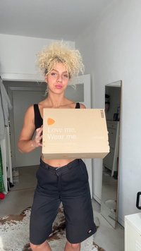 Boîte en carton marron avec le texte « Aime-moi. Porte-moi. », comportant un logo orange. Individu tenant la boîte dans une pièce aux murs clairs.